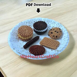 May include: A plate of crocheted biscuits and pastries. The plate includes a sandwich biscuit, a chocolate chip biscuit, a rectangular biscuit, a round biscuit, and a long, thin biscuit. The plate is on a light-coloured wooden surface. The text "PDF Download" is at the top.