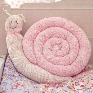 May include: A knitted snail-shaped pillow in soft pink and cream colors. The snail's shell is a spiral of pink, while the body is cream-colored. The snail has a smiling face and a pink bow. The pillow rests on a floral patterned fabric.
