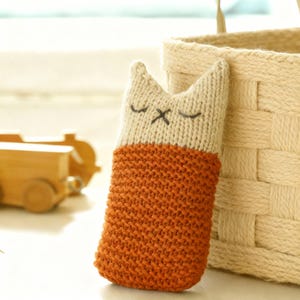 Puede incluir: Un juguete de peluche hecho a mano con forma de gato tejido. La parte superior es de color beige claro con ojos cerrados y una 'X' como boca. La parte inferior es de color naranja quemado. Una cesta tejida en el fondo. Aproximadamente 20 cm de alto.