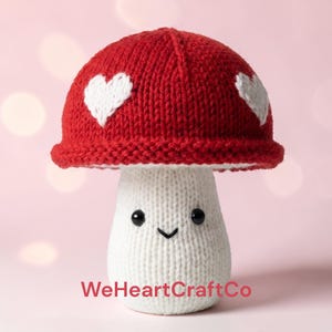 May include: A handmade knitted mushroom toy with a red cap featuring two white hearts. The mushroom's stem is white, with a smiling face and the text "WeHeartCraftCo" at the bottom. The toy is made of soft yarn.