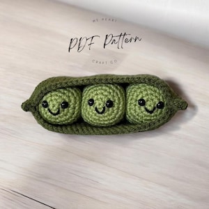 May include: A handmade crocheted pea pod containing three smiling peas. The peas are green with black eyes and smiles. The pod is also green. The text "PDF Pattern" and "Craft Co" are in the background.