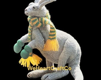 Peut inclure: Un kangourou gris tricoté portant une écharpe rayée verte et jaune et des mitaines assorties. Le kangourou a une longue queue et se tient debout. Le texte "WeHeartCraftCo" est visible en bas.