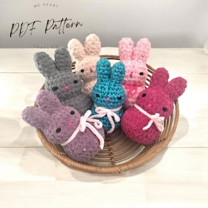 Puede incluir: Cinco peluches de conejo de crochet en una cesta de mimbre. Los conejos son en tonos de rosa, gris, morado, turquesa y burdeos. Cada conejo tiene una cinta rosa alrededor del cuello.