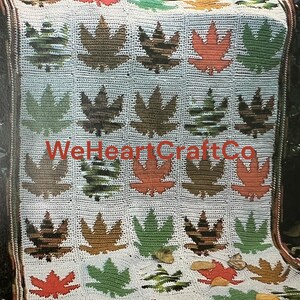 May include: A crocheted blanket featuring a repeating pattern of maple leaves in shades of brown, green, and orange on a white background. The blanket has a brown border and the text "WeHeartCraftCo" is visible.
