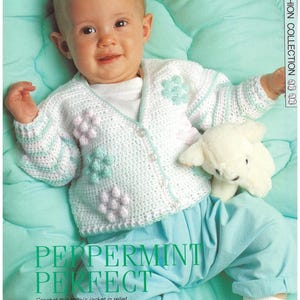 May include: A baby in a white crochet jacket with flower details and light blue trousers. The jacket features stripes and button fastenings. A soft toy is beside the baby. The image text says "PEPPERMINT PERFECT".