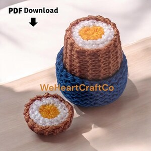May include: Handmade crochet sushi-themed items. The main piece is a brown and white sushi roll with a yellow center, sitting on a blue base. A smaller sushi roll is also present. The text "PDF Download" and "WeHeartCraftCo" are visible.
