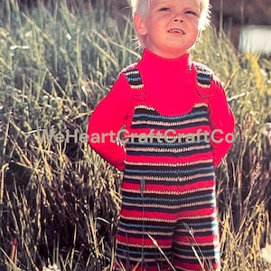 Op de afbeelding: Een kind in een rode coltrui en een gestreepte, gehaakte jumpsuit met rode, zwarte, blauwe en crèmekleurige strepen. Het kind staat in hoog gras in een zonnige buitenomgeving.