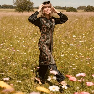 May include: A woman in a field of wildflowers wears a black outfit with a patterned vest. The long-sleeved top and pants are black. The vest features a detailed design in red, brown, and white. A black bow sits atop the woman's head.