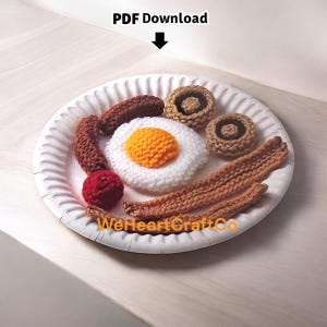 Puede incluir: Un plato de desayuno de ganchillo con un huevo frito, tocino, salchichas, un tomate y champiñones. El huevo tiene una yema amarilla y blanco, el tocino es marrón y la salchicha es de color marrón oscuro. El plato es blanco y tiene el texto "PDF Download" y "WeHeartCraftCo".