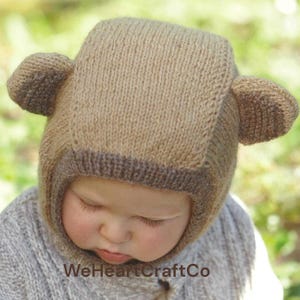May include: A knitted baby hat in shades of brown, designed to resemble a bear. The hat features ear-shaped protrusions on the sides and a darker brown trim around the face opening. The baby is wearing a grey sweater.
