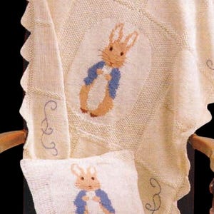 Könnte beinhalten: Cremefarbenes Strick-Babydecken- und Kissenset mit Peter Rabbit-Design. Die Decke hat einen gewellten Rand und ein zentrales Oval mit dem Hasen. Das Kissen hat das gleiche Design. Der Text "WeHeartCraftCo" ist sichtbar.