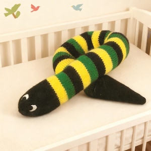 May include: A knitted snake toy in a cot. The toy has black, yellow, and green stripes. The head is black with white eye details. The tail is a solid black cone shape. Decorative bird cutouts are on the wall behind the cot.