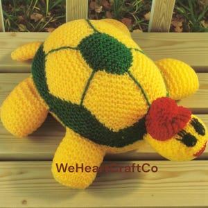 Puede incluir: Un peluche de tortuga tejido a mano en amarillo y verde, con un lazo en forma de corazón rojo. La tortuga tiene una cara sonriente. El texto "WeHeartCraftCo" es visible en la superficie de madera.