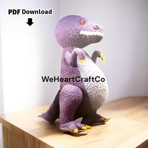 May include: A purple dinosaur plush toy with a white belly, red eyes, and yellow claws. The text "PDF Download" and "WeHeartCraftCo" are visible. The toy is sitting on a wooden surface.