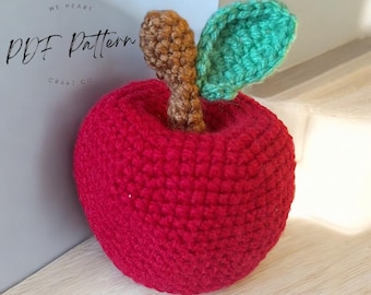 May include: A crocheted red apple with a brown stem and a green leaf. The apple is sitting on a wooden surface. The text "PDF Pattern" is visible in the background.