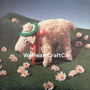May include: A whimsical, handmade sheep sculpture crafted from cream-colored yarn. It wears a green and white hat, a red ribbon, and has black eyes. The sheep stands on a green surface with scattered flowers. The text "WeHeartCraftCo" is visible.