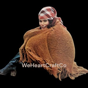 May include: A woman wearing a brown knitted shawl with fringe, a plaid headscarf, and black boots. The shawl is draped over the shoulders and around the body. The text "WeHeartCraftCo" is visible.