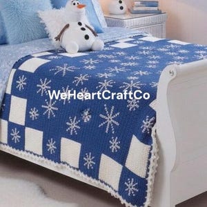 Op de afbeelding: Een blauw-witte gebreide deken met een sneeuwvlokkenpatroon. De deken heeft een dambordpatroon met witte en blauwe vierkanten, elk met witte sneeuwvlokken. De tekst "WeHeartCraftCo" is zichtbaar op de deken.