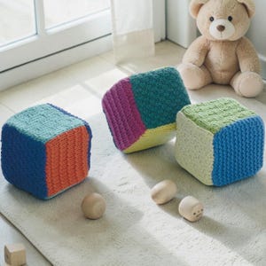 May include: Three colorful, knitted toy blocks in shades of blue, orange, green, pink, and yellow. The blocks are cube-shaped and sit on a light-colored rug. Two small wooden egg-shaped toys and a small wooden block are also present, along with a beige teddy bear.