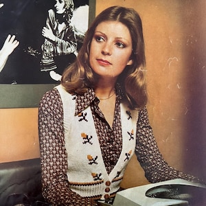 May include: A woman wearing a brown and white patterned blouse with a white knitted vest. The vest has a brown and white geometric pattern and buttons down the front. The woman is sitting at a desk with a typewriter in front of her.
