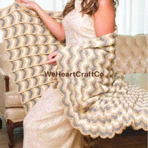 May include: A cream, grey, and tan chevron-patterned crochet shawl. The shawl has a scalloped edge and is draped over a person. The text "WeHeartCraftCo" is visible on the shawl.