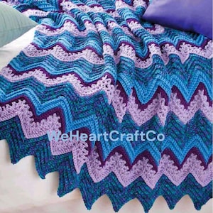 May include: A crocheted blanket featuring a zig-zag pattern in blue, teal, and purple hues. The blanket is displayed on a bed, highlighting its textured design. The text "WeHeartCraftCo" is visible on the blanket.