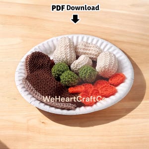 May include: A crocheted plate of food, including a brown item, white items, green balls, and orange shapes. The plate is white and the background is a light brown surface. The text "PDF Download" and "WeHeartCraftCo" are visible.