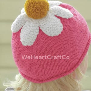 May include: A pink knit hat with a white daisy design and a yellow pom-pom center. The hat has the text "WeHeartCraftCo" on the side. The hat is a handmade item.