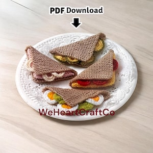 May include: Four crocheted sandwich halves are arranged on a white plate. The sandwiches are brown with fillings like egg, tomato, and lettuce. The text "PDF Download" and "WeHeartCraftCo" are visible.
