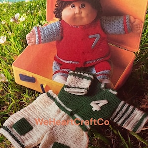 May include: A Cabbage Patch Kids doll wearing a red and blue crocheted outfit with the number 7. The doll is sitting in an orange suitcase. Two pairs of crocheted shorts, one white and green, the other green and white, are in the foreground. The text "WeHeartCraftCo" is visible.