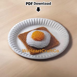 May include: A crocheted fried egg on toast, presented on a white paper plate. The egg features a white yolk and a bright orange yolk, atop a square of brown toast. The plate is on a light brown surface. Text on the image reads "PDF Download" and "WeHeartCraftCo".