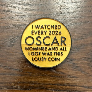 May include: A round, yellow coin with black text that reads "I WATCHED EVERY 2026 OSCAR NOMINEE AND ALL I GOT WAS THIS LOUSY COIN". The coin has a black border and is set against a wooden surface.