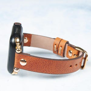 May include: A brown leather watch band with a gold buckle and gold-colored hardware. The watch face is black. The band is made of textured leather.
