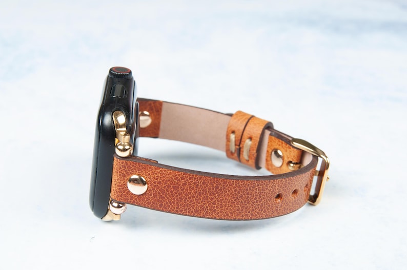 May include: A brown leather watch band with a gold buckle and gold-colored hardware. The watch face is black. The band is made of textured leather.