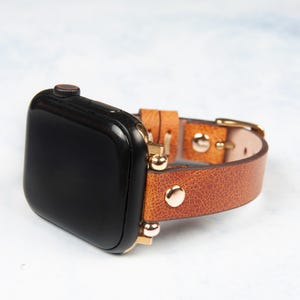 May include: A black smartwatch with a brown leather band. The band features gold-colored hardware and rivets. The watch is displayed on a light-colored surface.