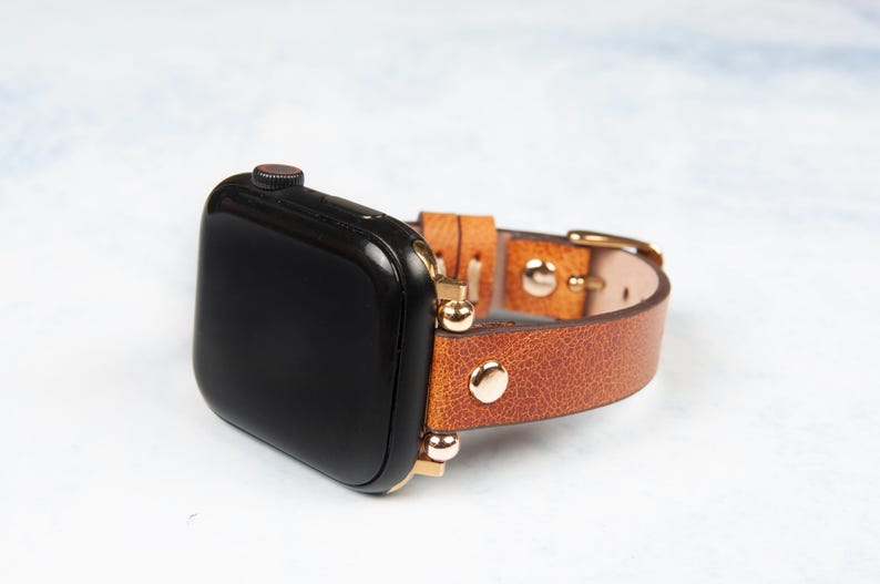 May include: A black smartwatch with a brown leather band. The band features gold-colored hardware and rivets. The watch is displayed on a light-colored surface.