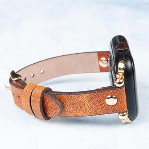 May include: A brown leather watch band with a black watch face. The band has a textured appearance and is secured with gold-colored hardware. The watch face has a red accent. The band is designed to fit a smartwatch.