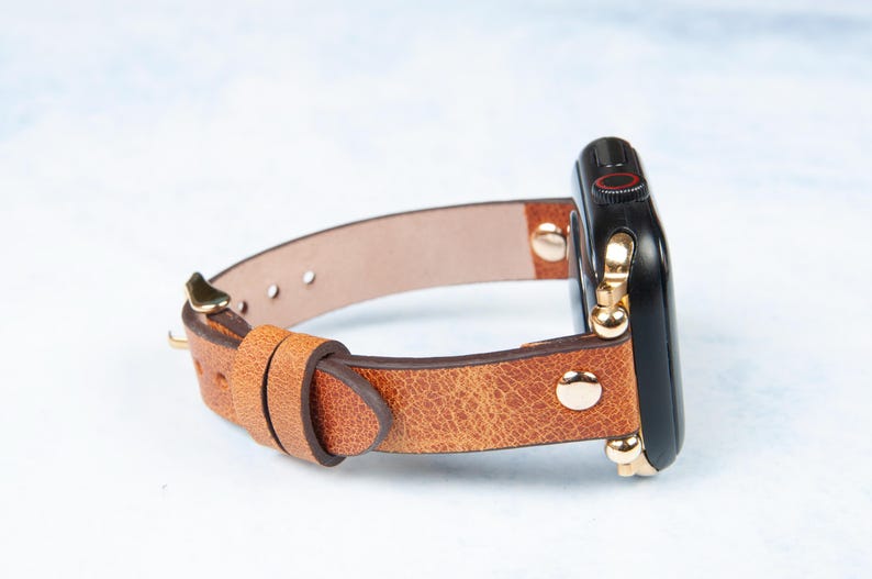 May include: A brown leather watch band with a black watch face. The band has a textured appearance and is secured with gold-colored hardware. The watch face has a red accent. The band is designed to fit a smartwatch.