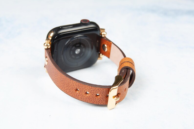 May include: A brown leather watch band with gold-colored accents and a gold buckle. The watch face is black. The band is made of a light brown leather and is secured with a gold-colored buckle.
