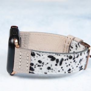 May include: Black smartwatch with a light beige leather strap and a cowhide patterned band. The watch band features a speckled black and white design, and the watch case has rose gold accents.