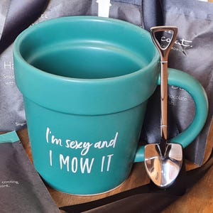 May include: A teal green ceramic mug shaped like a flower pot with the text "I'm sexy and I MOW IT" printed on the side. A silver metal spoon shaped like a small shovel is next to the mug.
