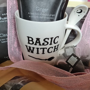 May include: A white mug with the text "BASIC WITCH" and a black broom graphic. The mug is surrounded by tea bags, a pink ribbon, and a white spoon with a crescent moon design.