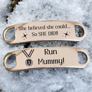 Könnte beinhalten: Zwei silberne Schuh-Anhänger mit eingraviertem Text. Der obere Anhänger lautet "She believed she could... So SHE DID!!" mit zwei Sternen. Der untere Anhänger lautet "✓ Run Mummy!" mit einer Medaillengrafik.