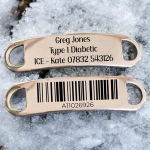 Op de afbeelding: Twee medische waarschuwingslabels in roségoud. Op het bovenste label staat "Greg Jones, Type 1 Diabeet, ICE - Kate 07832 543126." Het onderste label heeft een barcode en het nummer "A11026926."