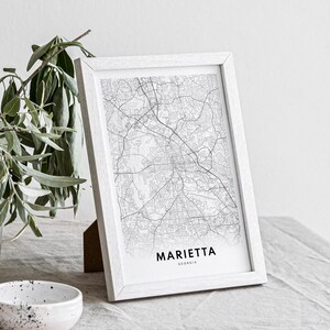 May include: A white framed print of a black and white map of Marietta, Georgia. The print is on a white surface with a green plant in the background.