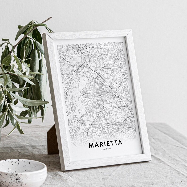 May include: A white framed print of a black and white map of Marietta, Georgia. The print is on a white surface with a green plant in the background.