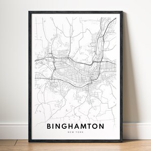 Op de afbeelding: Een ingelijste zwart-wit kaartafdruk van Binghamton, New York. De kaart toont de straten, wegen en waterwegen van de stad. De naam van de stad is gedrukt in een vetgedrukte, schreefloze lettertype onderaan de afdruk, met "New York" eronder.