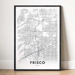Op de afbeelding: Een ingelijste zwart-wit kaartprint van Frisco, Texas. De kaart toont de straten en infrastructuur van de stad. De naam van de stad en de staat zijn onderaan in een vetgedrukte, schreefloze lettertype gedrukt.