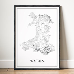 May include: A black and white line art map of Wales with the word "WALES" printed below the map. The map is framed in a black frame and is hanging on a white wall.