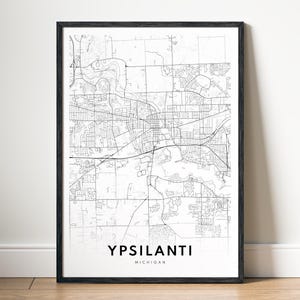 May include: A framed black and white map print of Ypsilanti, Michigan. The map shows the city's streets and infrastructure. The city name and state are printed in a clean, modern font at the bottom of the print.